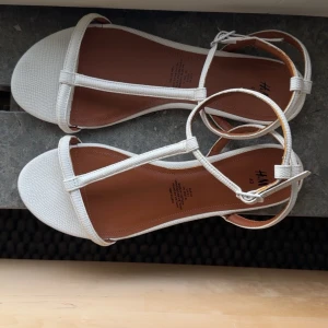 Vita REMSANDALER från H&M - Säljer ett par stilrena sandaler från H&M i storlek 40 i samma modell som bild 2 och 3. De har en platt sula, rund tå och smala remmar med snörning runt ankeln. Färgen är vit och materialet är syntet. 