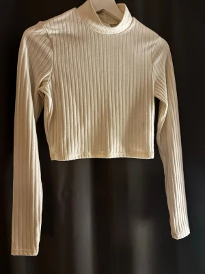 Ribbad croppad polotopp i beige - Säljer en beige croppad polotopp med lång ärm och ribbad struktur. Toppen har en hög krage och är tillverkad i ett mjukt material som ger en skön känsla mot huden. Perfekt att styla med jeans eller kjol för en clean look.