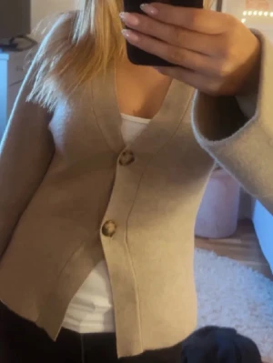 Beige kofta från Gina Tricot - Säljer en beige kofta från Gina Tricot med v-ringning och stora dekorativa knappar framtill. Koftan är stickad, har lång ärm med uppvik och knappdetalj vid ärmslut. Perfekt till lager på lager och enkel att styla.