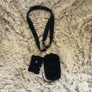 yaya phone bag från Gina Tricot - En mobil väska och AirPod väska från Gina Tricot.  Svart axelväska med bred justerbar rem och två separata fack. Ena facket har dragkedja och elastisk snörning på framsidan, det andra har tryckknapp. Perfekt för att bära mobil, kort och småprylar. Enkel och stilren design.