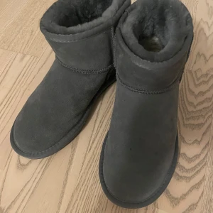 Mörkgrå UGG boots i fårskinn - Säljer ett par mörkgrå UGG boots i mjukt fårskinn med fluffigt foder och platt sula. Klassisk rund tå och robust yttersula med bra grepp. Perfekta för kalla dagar och riktigt sköna att dra på sig när det är kyligt ute. Tyvärr ingen låda. 