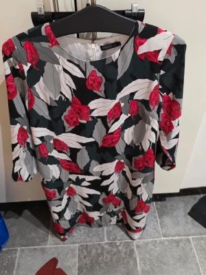 Blommig klänning från STOCKH LM - Snygg kortklänning med blommigt mönster i rött, vitt, grått och svart från STOCKH LM. Klänningen har rund hals, trekvartsärm och dragkedja i ryggen. Materialet är mjukt och känns som polyester, perfekt för dig som gillar färgstarka prints.