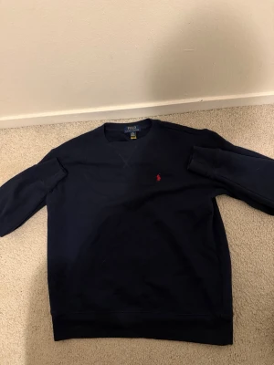Mörkblå sweatshirt från Polo Ralph Lauren - Snygg mörkblå sweatshirt från Polo Ralph Lauren med klassisk rund hals och långärmad design. Tröjan har en liten röd broderad logga på bröstet och är gjord i mjuk bomull. Perfekt för chill dagar och enkel att matcha med jeans eller joggers. Passar dig som är cirka 160 cm lång.