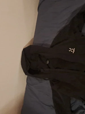 Svart vindjacka från Haglöfs med huva - Svart vindjacka från Haglöfs med dragkedja och huva. Jackan har en stilren look med logga på bröstet och praktiska fickor med dragkedja. Perfekt för blåsiga dagar och outdoor-aktiviteter. Snygg och funktionell design.