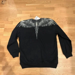 Svart tröja med vingeprint Marcelo Burlon - Svart sweatshirt från Marcelo Burlon County of Milan med ikoniskt vingeprint över axlar och bröst. Tröjan har rund hals, långa ärmar och ribbade muddar. Perfekt statement piece för dig som vill sticka ut med en edgy look. Oanvänd och riktigt fräsch 