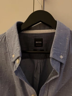 Blå skjorta från BOSS med mönster - Snygg blå skjorta från BOSS med diskret broderat mönster och klassisk krage. Skjortan har vita knappar och lång ärm, perfekt för dig som gillar stilrena detaljer och en modern passform. XL passar även L. 