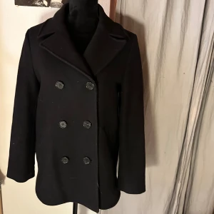 Svart skepparkavaj från & Other Stories - Klassisk svart skepparkavaj från & Other Stories med dubbelknäppning och bred krage. Jackan har en stilren och tidlös look, perfekt för dig som gillar minimalistisk stil. Tillverkad i ullblandning och har långa ärmar samt snygga knappar framtill.