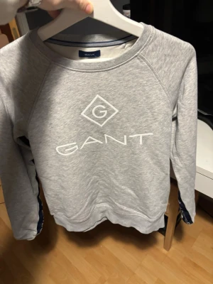 Grå sweatshirt från GANT - Säljer en stilren grå sweatshirt från GANT med stor vit logga framtill. Tröjan har rund halsringning och långa ärmar. Tillverkad i mjuk bomullsblandning som är perfekt för både skola och chill. Klassisk passform och enkel design.