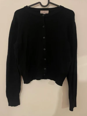 Svart kofta från H&M med knappar - Svart, klassisk kofta från H&M i storlek M. Koftan har rund halsringning, långa ärmar och knäppning med svarta knappar framtill. Materialet är mjukt och stickat, perfekt för lager-på-lager under kyliga dagar. Passar XS likt S 