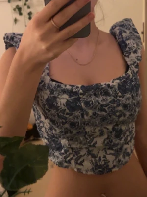 Blommig blåvit korsettopp - Supersöt korsettopp med blått blommönster på vit botten. Toppen har breda axelband och fyrkantig urringning som ger en romantisk vibe. Croppad modell som passar perfekt till höga jeans eller kjol. Materialet känns stadigt och håller formen snyggt. 
