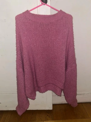 Rosa stickad tröja Meet Me There - Mysig rosa stickad tröja från Meet Me There med loose passform och bred ribbad kant vid halsen, ärmslut och nederkant. Tröjan har långärmade, lite oversized ärmar och är perfekt för kyliga dagar. Färgen är en mjuk rosa och materialet känns fluffigt och skönt.