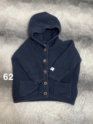 Mörkblå stickad kofta med huva - Mysig mörkblå stickad kofta med huva och träknappar framtill. Koftan har två fickor fram och en grovstickad struktur som ger en cool look. Perfekt för kyliga dagar och enkel att matcha med andra plagg.🌸 Fint använt skick. Storlek 62