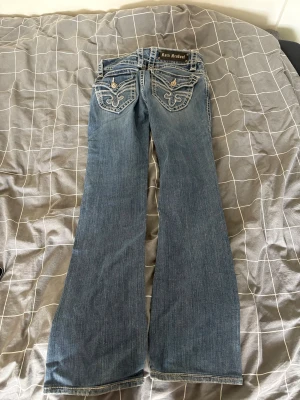 Rock Revival blå  jeans - Säljer ett par blå jeans från Rock Revival med tydliga kontrastsömmar och broderade bakfickor med metallknappar. Klassisk bootcut -modell med låg midja och colla detaljer . 