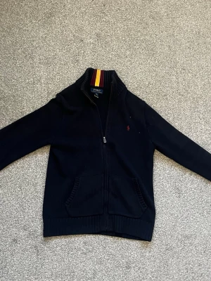 Svart stickad kofta från Polo Ralph Lauren - Svart stickad kofta från Polo Ralph Lauren med dragkedja framtill och ribbad krage med röd, gul och vinröd rand. Klassisk logga broderad på bröstet och två fickor fram. Perfekt för dig som gillar stilrena kläder. Kan diskutera pris!