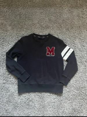 Svart sweatshirt Morris R. Club S - Svart sweatshirt från Morris R. Club i storlek S. Tröjan har ett stort rött M-tryck på bröstet och vita ränder på ena ärmen. Rund halsringning med ribbad kant och mjukt bomullsmaterial. Perfekt för en chill och sportig look.