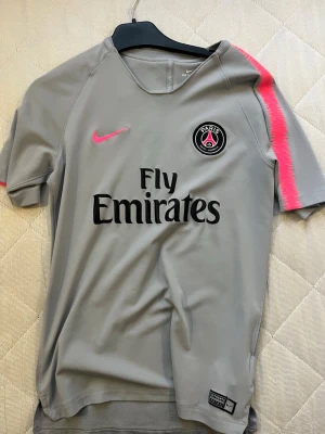 Grå PSG Nike fotbollströja - Säljer en original  grå Paris Saint-Germain fotbollströja från Nike med rosa detaljer på axlarna och rosa Nike-logga. Tröjan har Fly Emirates-tryck på bröstet och PSG-emblem. Materialet är lätt och andas, perfekt för fotboll eller träning.