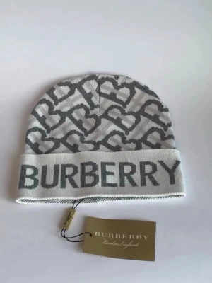 Gråvit Burberry mössa med hjärtan - Snygg mössa från Burberry i grått och vitt med stickat hjärtmönster och stor logotyp framtill. Mössan är tillverkad i mjukt stickat material och har en bred uppvikt kant. Perfekt för dig som vill ha en cool accessoar med lyxig känsla.