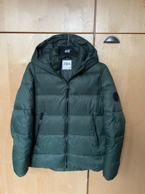 Grön pufferjacka från Zara XS - Snygg mörkgrön pufferjacka från Zara i storlek XS. Jackan har huva, två sidofickor med dragkedja och en svart patch på ärmen. Materialet är syntet och jackan är thermo-isolerad samt vattenavvisande. Perfekt för kalla dagar.💗