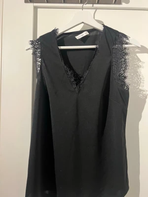 Svart spetsdetaljerad blus från Gallery - Svart blus från Gallery med v-ringning och snygga spetsdetaljer längs halsringning och axlar som är ärmlös. Använd 2 gånger.