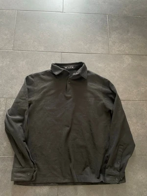 Svart långärmad henley från Zara - Svart henley-skjorta från Zara med klassisk krage och två knappar vid halsen. Skjortan är långärmad och har en enkel, stilren design utan mönster. Materialet är mjukt och känns som bomull, perfekt för en chill och clean look.