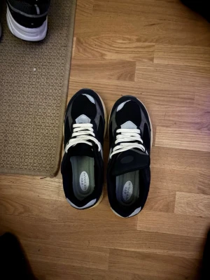 New Balance 2002r svarta sneakers - Säljer ett par New Balance 2002r sneakers i svart med grå och vita detaljer. Skorna har mesh och mocka på ovandelen, vit snörning och en beige platt sula. Reflekterande inslag på tån och sidan samt den klassiska N-loggan. Perfekt för dig som gillar stilrena och bekväma sneakers.