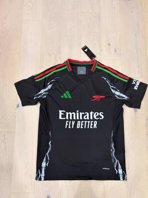 Arsenal x Adidas svart fotbollströja M - Svart Arsenal fotbollströja från Adidas med gröna och röda ränder på axlarna, broderat rött kanonmärke och vit Emirates-tryck på bröstet. Tröjan har mönstrade detaljer i grått och är tillverkad i polyester med Aeroready-teknologi. Slim fit-modell.