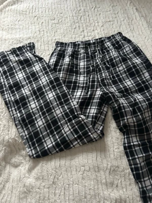 Svarta och vita rutiga pyjamasbyxor  - Säljer ett par pyjamasbyxor med svart och vitt rutmönster. De har elastisk midja med snörning och är tillverkade i mjukt bomullsmaterial. Passformen är loose och benen är raka för en chill vibe. Perfekta för att hänga hemma eller bara vara bekväm.