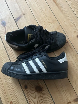 Adidas Superstar svarta sneakers - Klassiska Svarta Adidas Superstar sneakers med vita ränder på sidorna. Skorna har använts men inte det senaste året.