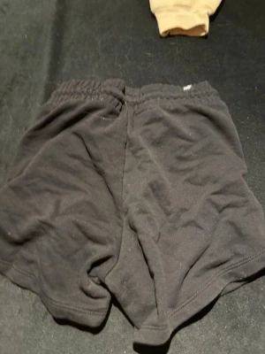 Svarta shorts med resår och snörning - Svarta shorts från Puma med elastisk midja och snörning framtill. Mjukt bomullsmaterial och klassisk passform. Puma-logga i vitt på ena benet ger en sportig vibe. Perfekta för chill eller träning.