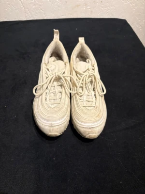 Nike Air Max 97 vita sneakers - Nike Air Max 97 sneakers i helvitt med ikoniska vågiga linjer och synlig Air-sula längs hela skon. Skorna har snörning och en platt sula med rund tå. Materialet är syntet och skinn, och de har en sportig vibe som passar perfekt till streetwear.