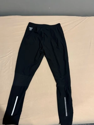 Svarta träningsbyxor med reflexdetaljer - Säljer ett par svarta träningsbyxor med smal passform och elastisk midja. Byxorna har reflexdetaljer på benen och dragkedja vid bensluten, perfekta för löpning eller annan träning. Materialet är lätt och syntetiskt för bra ventilation.
