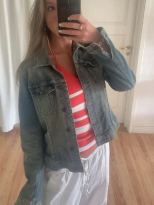 Klassisk blå jeansjacka - Säljer en klassisk blå jeansjacka med knappar framtill och två bröstfickor. Jackan har en tidlös design med krage och är perfekt att slänga över en t-shirt eller hoodie. Materialet är slitstarkt denim och jackan har en normal passform med långa ärmar.