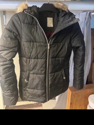 Svart pufferjacka från Etirel med fuskpäls - Svart pufferjacka från Etirel i storlek 34. Jackan har huva med beige fuskpäls, quiltad design och dragkedja framtill. Fickor med knappdetaljer och snygga sömmar ger en cool look. Perfekt för kalla dagar och har en normal passform.