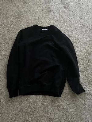 Svart sweatshirt från Lacoste - Svart sweatshirt från Lacoste i regular fit med klassisk rund hals och broderad blå krokodil på bröstet. Tröjan har långa ärmar och ribbade muddar vid hals, ärmslut och nederkant. Perfekt för en chill och stilren look. Säljer billigt för behöver pengar