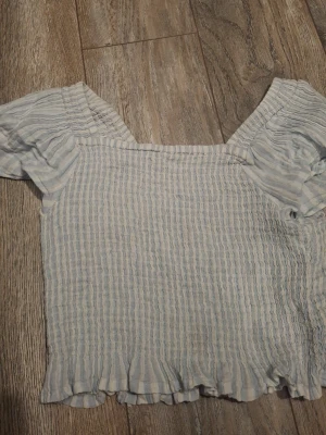 Randig blus från Kappahl - Söt blus från Kappahl i ljusblått och vitt med smala ränder. Modellen har korta puffärmar, fyrkantig ringning och volangkant nertill. Tyget är lätt och stretchigt, perfekt för varma dagar.