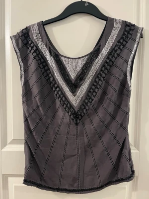 Grå paljettblus från H&M - Säljer en grå blus från H&M i storlek 38. Toppen har V-ringning med silverfärgade paljetter och svarta broderade detaljer som bildar ett snyggt mönster framtill. Ärmlös och tillverkad i ett lätt, syntetiskt material. Perfekt för dig som gillar lite extra bling.