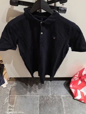 Svart pikétröja från GANT - Säljer en klassisk svart pikétröja från GANT med korta ärmar och krage. Tröjan har två vita knappar framtill och en liten broderad GANT-logga på bröstet. Materialet är mjuk bomull som andas och passar perfekt till en chill stil.