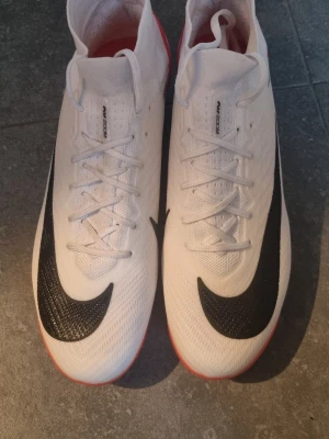 Nike Zoom fotbollsskor vit/röd - Säljer ett par Nike Zoom fotbollsskor i vitt med röda och svarta detaljer. Skorna har snörning, svart Nike-logga på sidan och en sula med röda dobbar för bra grepp på planen. Ovandelen är i syntetmaterial och designen är riktigt sportig.