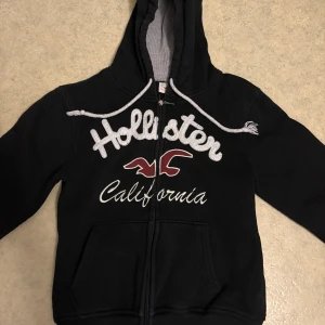 Svart Hollister hoodie med dragkedja - Svart hoodie från Hollister med stor broderad logga och text på bröstet. Tröjan har huva med grått foder, vita snören och praktiska fickor framtill. Stängs med dragkedja och har långa ärmar. Perfekt för chill dagar och streetstyle. Storlek M men passar S