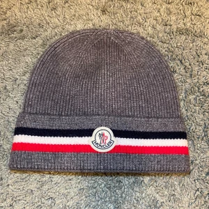 Moncler mössa i grå - Snygg grå ribbstickad mössa från Moncler med klassiska ränder i marinblått, vitt och rött längs kanten. Framtill sitter Monclers ikoniska logotyp. Perfekt för dig som vill ha en stilren och exklusiv look under kyliga dagar. Den är helt oanvänd och med tags kvar och qr koden funkar.