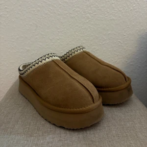 Beige mocka slip-in sandaler med plattform - Säljer ett par beige slip-in sandaler med plattformsula och rund tå. Skorna är tillverkade i mjuk mocka och har en dekorativ kant med svartvitt zigzag-mönster. Perfekta för chill dagar och har en cozy vibe.