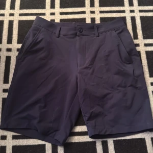Mörkblå shorts från Under Armour - Snygga mörkblå shorts från Under Armour med klassisk design och raka ben. De har fickor fram och bak, bälteshällor och knappstängning. Materialet är mjukt och lätt polyester, perfekt för en chill sommardag. Diskret logga bak ger en sportig touch.