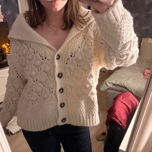 Krämvit stickad kofta med knappar - Supermysig krämvit stickad kofta med grovt mönster och stora knappar framtill. Koftan har en bred krage och långärmade ärmar med ribbade muddar