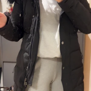 Svart lång dunjacka med huva Hollie’s  - Säljer en svart dunjacka med huva och snyggt pälskant. Jackan är lång, har dragkedja och knappar framtill samt praktiska fickor på sidorna. Perfekt för kalla dagar och har en stilren look som funkar till allt.