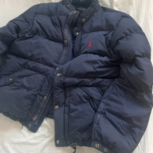 Mörkblå pufferjacka från Polo Ralph Lauren - Säljer en mörkblå pufferjacka från Polo Ralph Lauren med röd broderad logga på bröstet. Jackan har dragkedja, knappar och snörning nertill för extra passform. Perfekt för kalla dagar. .