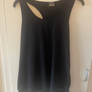 Svart linnetopp från Newbody - En svart, stilren linnetopp från Newbody med klassisk rund halsringning och ärmlös design. Tillverkad i mjuk polyesterblandning med stretch för en bekväm passform. Perfekt för träning eller som basplagg till din garderob.