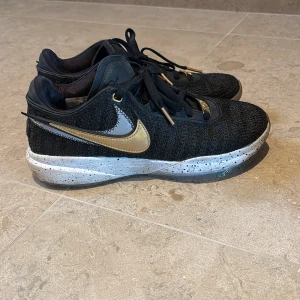 Svarta Nike sneakers med gulddetaljer - Snygga svarta Nike sneakers med stickad ovandel och guldfärgade detaljer på snörningen och swoosh-loggan. Skorna har en vit, stänkmönstrad mellansula och genomskinlig yttersula med gröna inslag. Perfekt för dig som gillar sportig och unik stil.