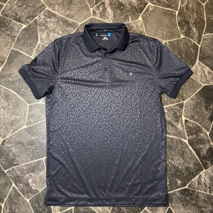 J.Lindeberg Piké - Snygg mörkblå pikétröja från J.Lindeberg med diskret geometriskt mönster över hela plagget. Slim fit-passform, klassisk krage och tre knappar framtill. Tillverkad i mjuk polyester som andas. Perfekt för dig som gillar stilrena detaljer och sportig vibe.