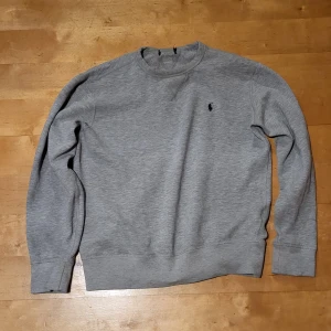 Grå sweatshirt från Polo Ralph Lauren - Snygg grå sweatshirt från Polo Ralph Lauren med klassisk rund hals och broderad logga på bröstet. Tröjan har långärmad passform och är tillverkad i mjuk bomull, perfekt för chill dagar. Enkel och stilren design som funkar till jeans eller joggers.