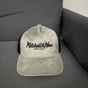 Mitchell & Ness grå och svart keps - Snygg keps från Mitchell & Ness med grå front i mjuk sammet och svart mesh på sidorna och bak. Broderad logga framtill och justerbar snapback. Perfekt för dig som gillar streetwear och vill ha en unik look.